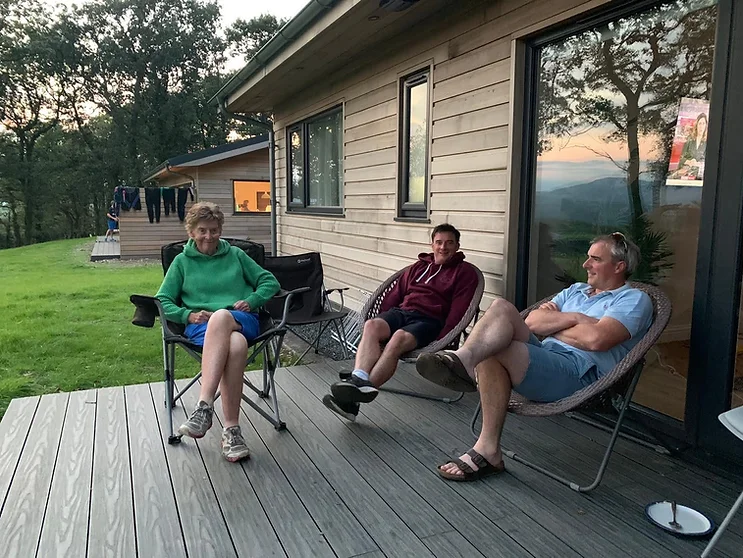 Three individuals stand on the lodge's decking, overlooking serene natural surroundings enjoying haven of relaxation and luxury, promising unforgettable moments amidst breath taking scenery